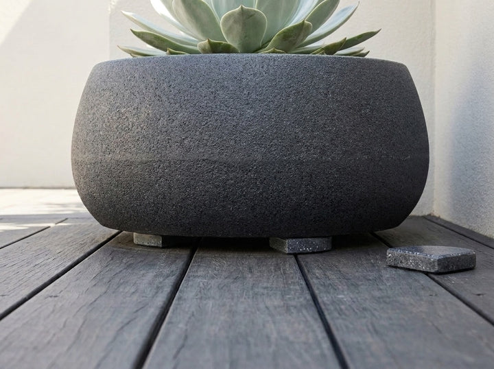 A Onyx deep near-black terrazzo planter lifted clear of the paving on Onyx pot feet, with one loose foot beside it - each foot is 70mm across and raises the pot 15mm for drainage and airflow