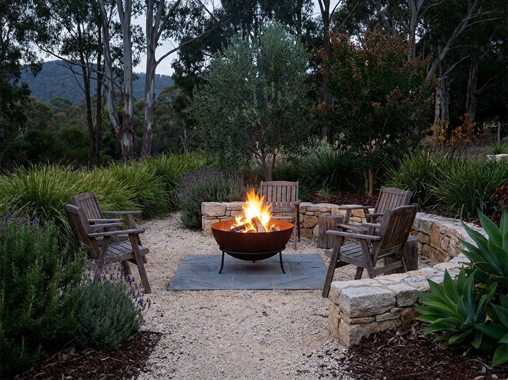 Steel Fire Pit, 61 cm across and 43.5 cm high, burning in a gravel fire-pit clearing in an Adelaide Hills garden at dusk with chairs drawn up around it